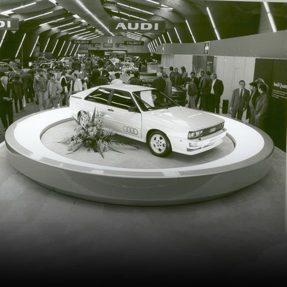 Three-quarter side profile of an early Audi racing vehicle parked on a pedestal with people looking on in the background.