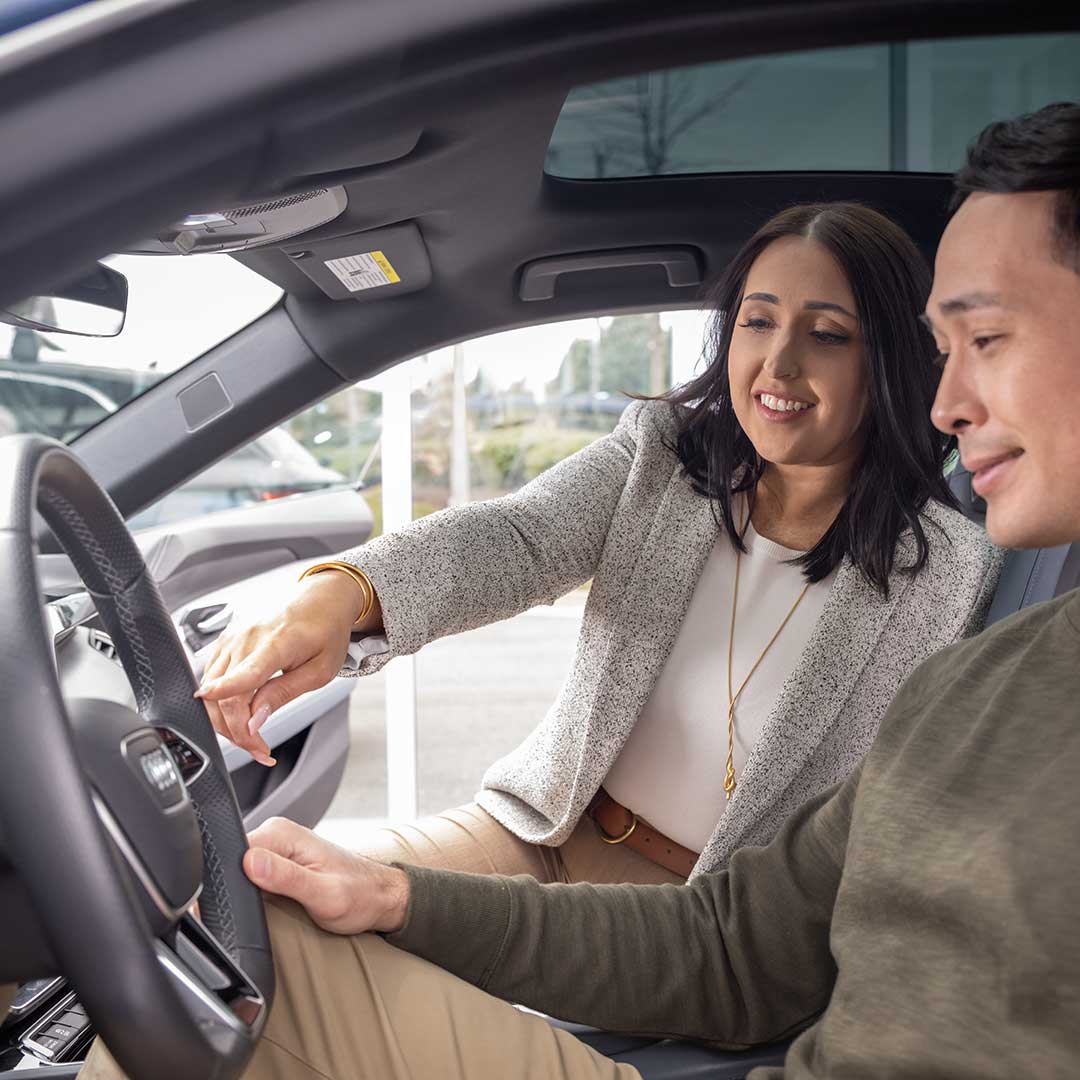 An Audi dealer explaining the features of an Audi vehicle steering wheel to a customer in the driver's seat.