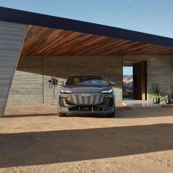 Front profile of a grey Audi Q6 Sportback e-tron parked and charging in front of a modern home.