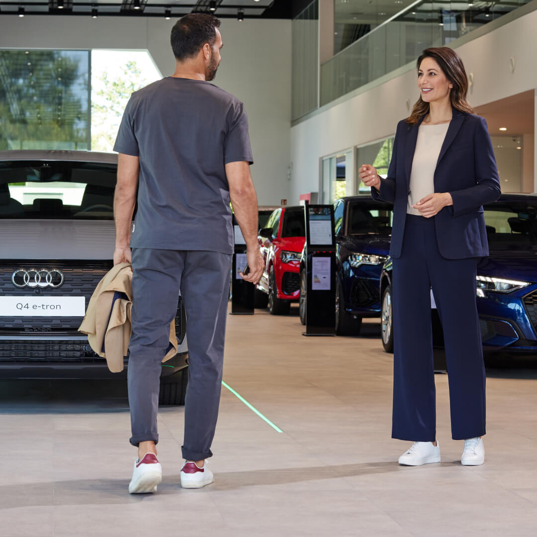 Audi salesperson speaks with a customer beside Audi vehicles in a bright dealership showroom.