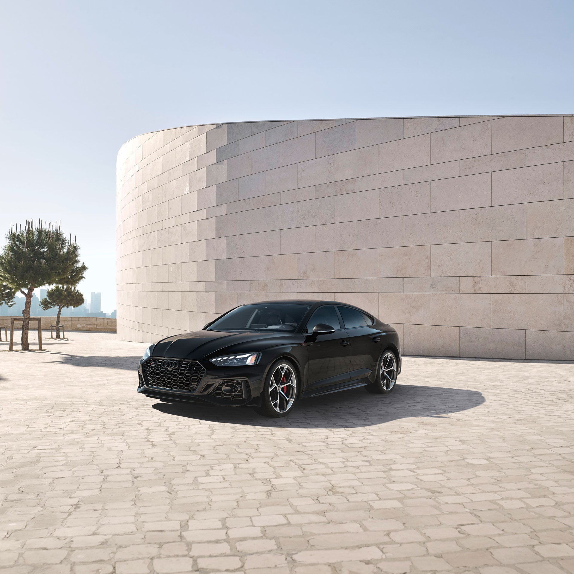 A black Audi RS 5 Coupe parked on a tiled road and tiled wall, near a few planted trees.