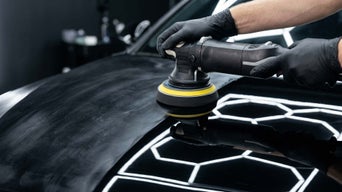Close-up of a technician polishing a black vehicle hood with a rotary tool.