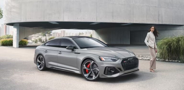 A gray Audi sports sedan parked outside a modern building with a woman standing nearby.