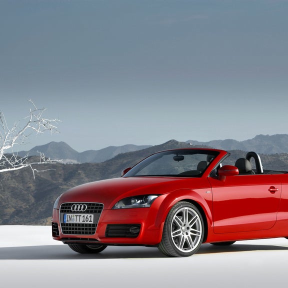 A red TT Coupe parked on snowy ground next to a tree in front of mountains.