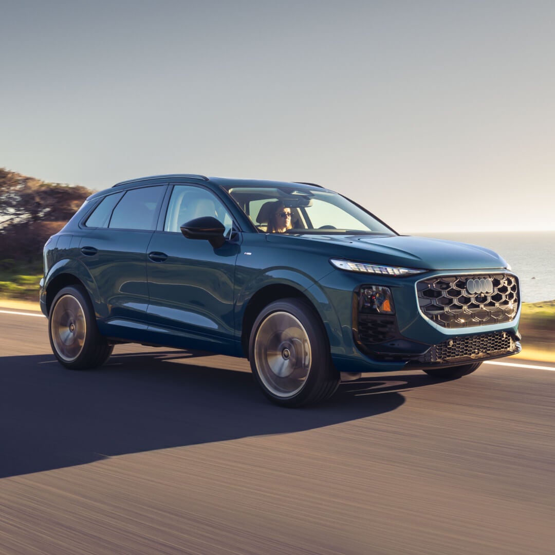 Side exterior action shot of a blue 2027 Audi Q3 driving on a seaside highway.