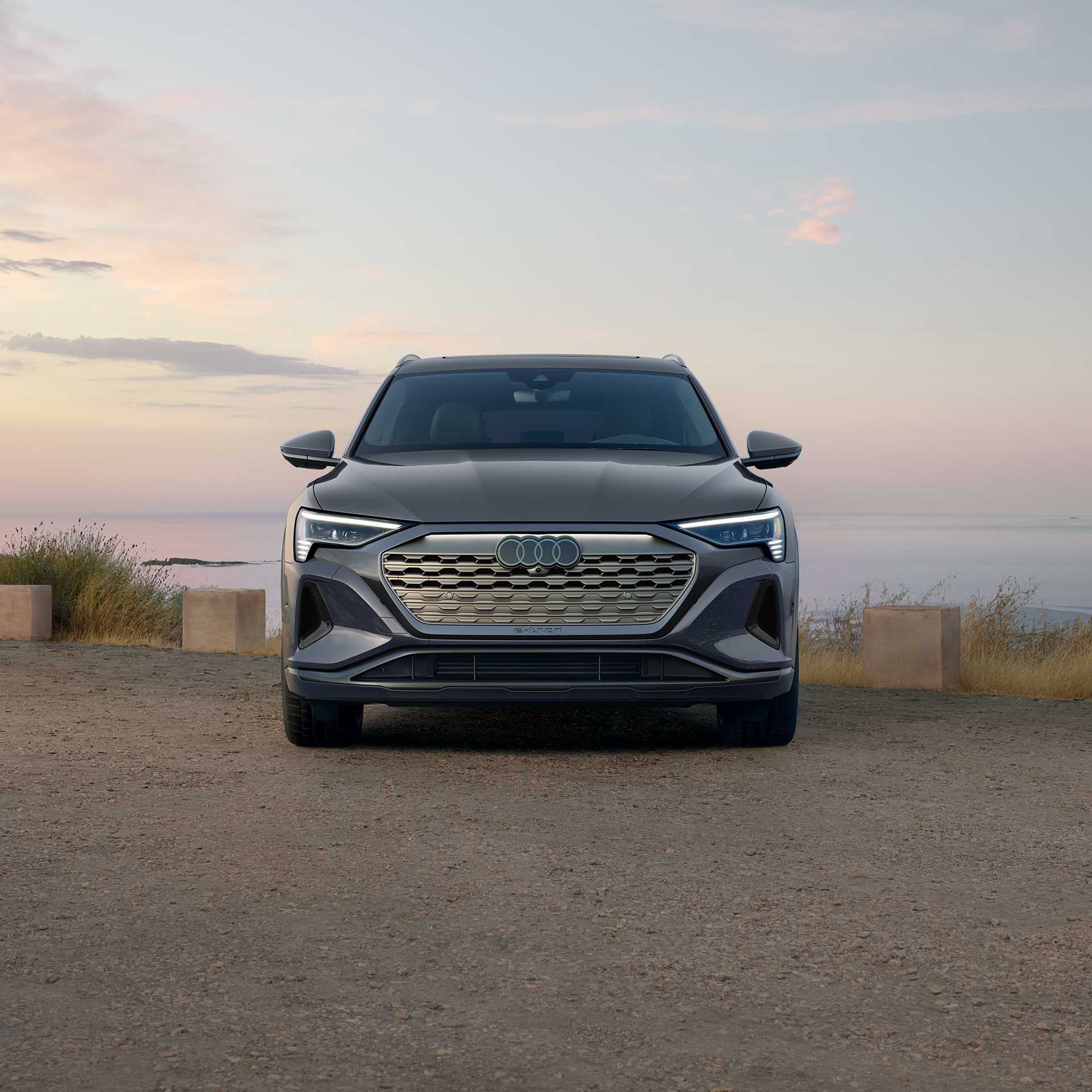 Front view of a gray Audi Q8 e-tron parked on a patch of dirt next to a body of water. 