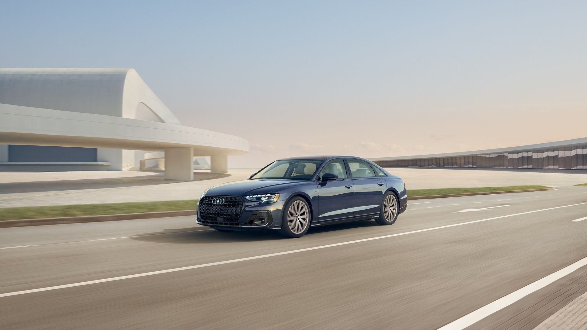 Blue A8 driving down a concrete road with a park strip and a modern building in the background. 