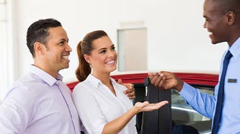 Three people standing in a dealership as one hands over car keys beside a red car.