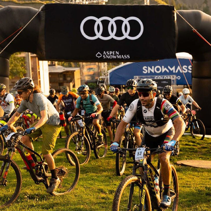 A group of bikers starting a race with an Audi Aspen Snowmass banner in the background.