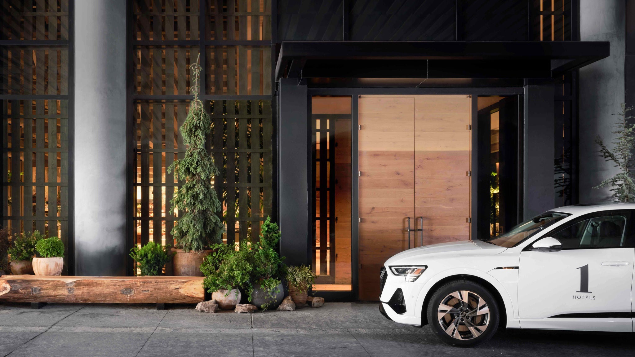 Side profile of a white Audi e-tron with a 1 Hotels logo on it parked in front of an upscale hotel. 