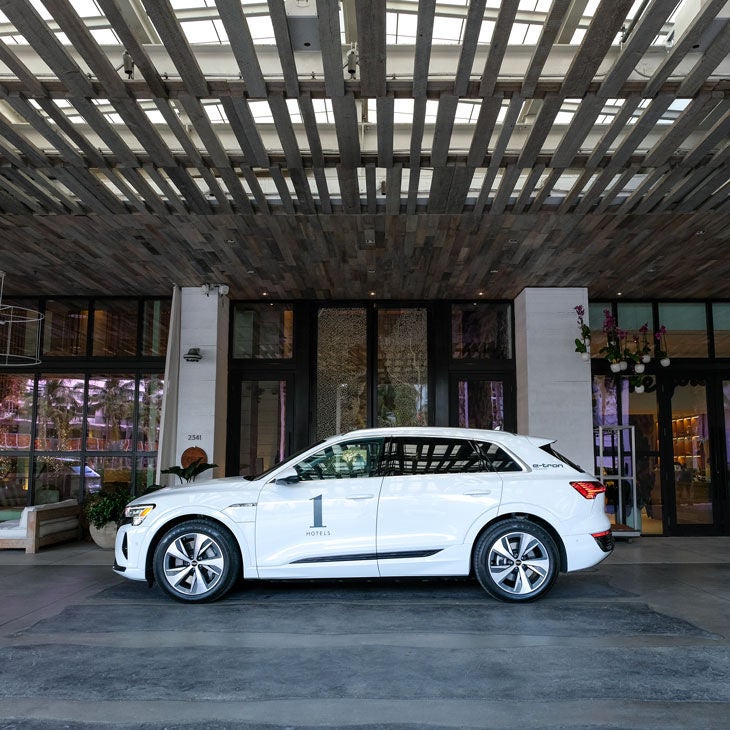 Side profile of a white Audi e-tron vehicle parked in front of an 1 Hotel location entrance.