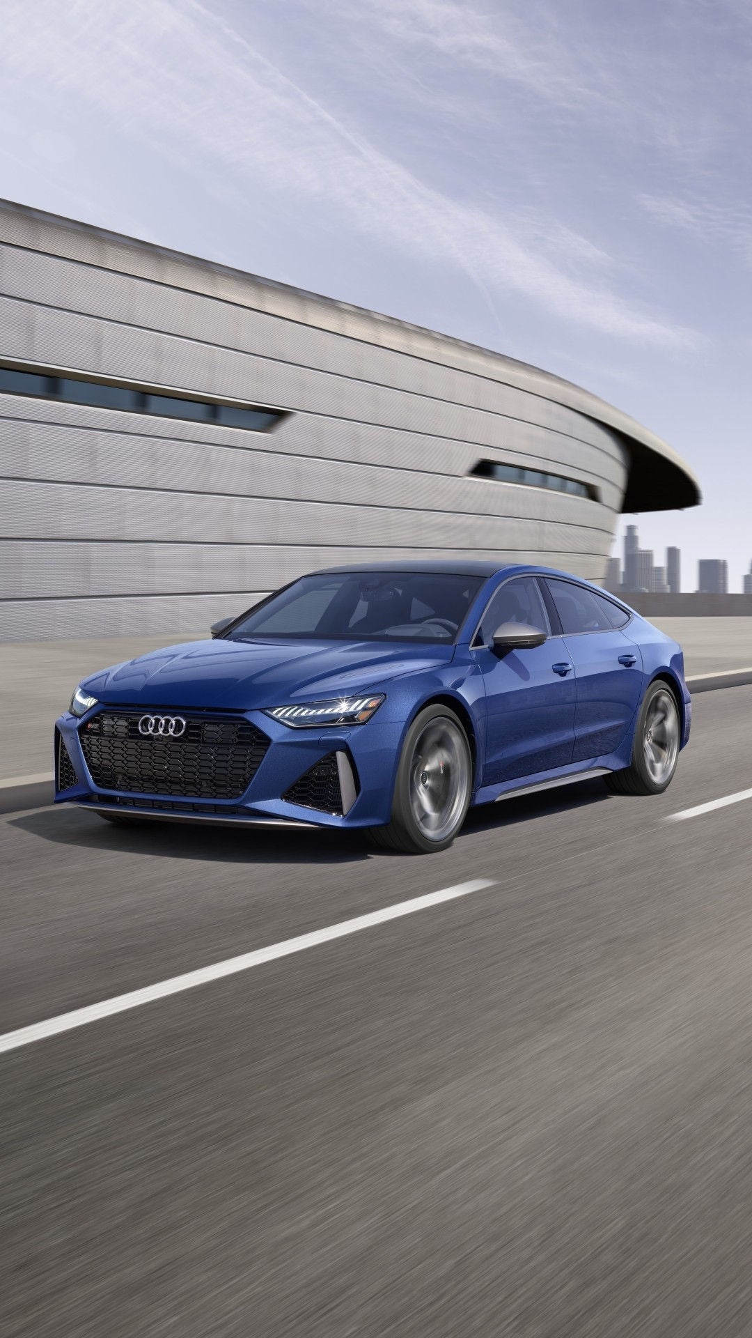 3/4 front view of Audi RS 7 passing a modern building on a city road.