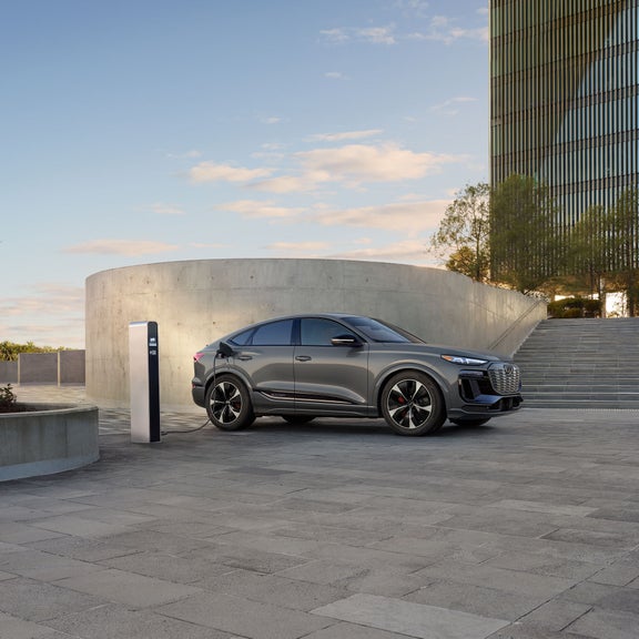 Vue latérale d'une Audi SQ6 Sportback e-tron grise garée et en charge près d'un bâtiment moderne.
