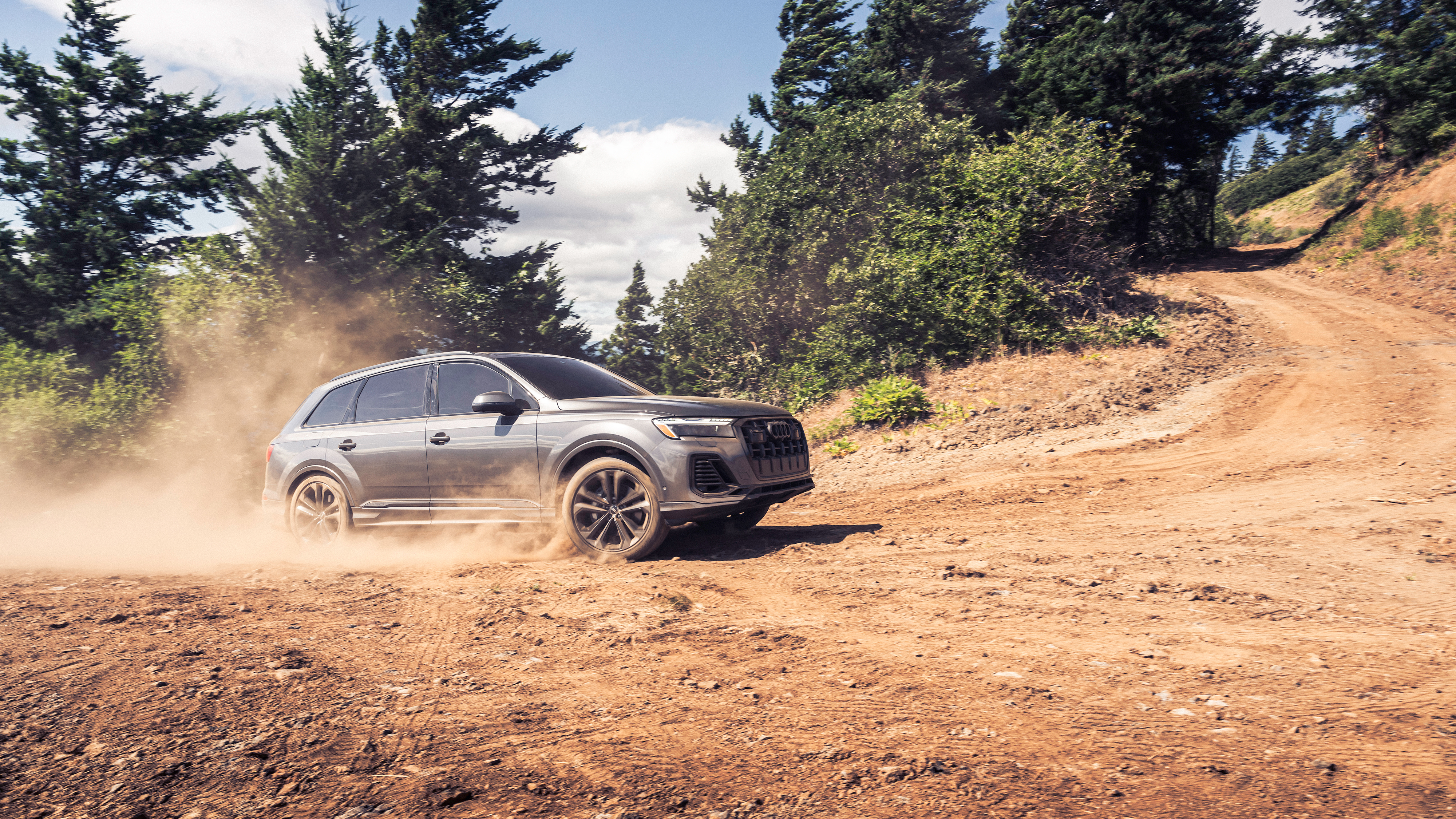 Gray Audi 2026 Q7 driving on a dirt ground with a background of trees. 