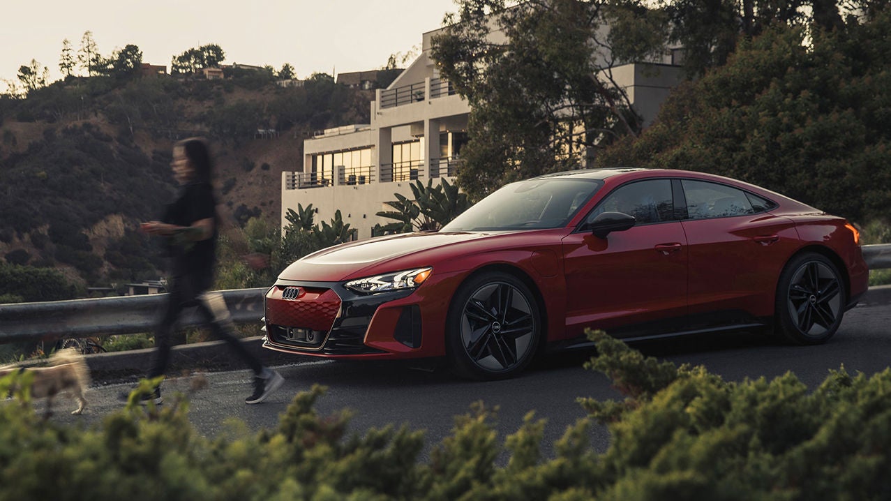 An Audi e-tron vehicle driving on a road, near a woman walking her dog, a house and a hill crowned with other houses.