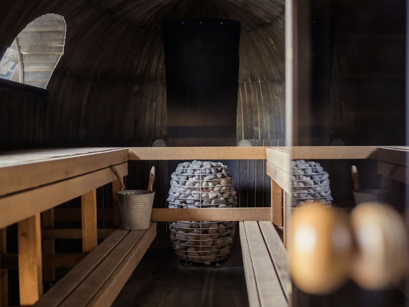 Inside view of a circular wooden sauna cabin with a central cage-style stone heater and sauna bucket