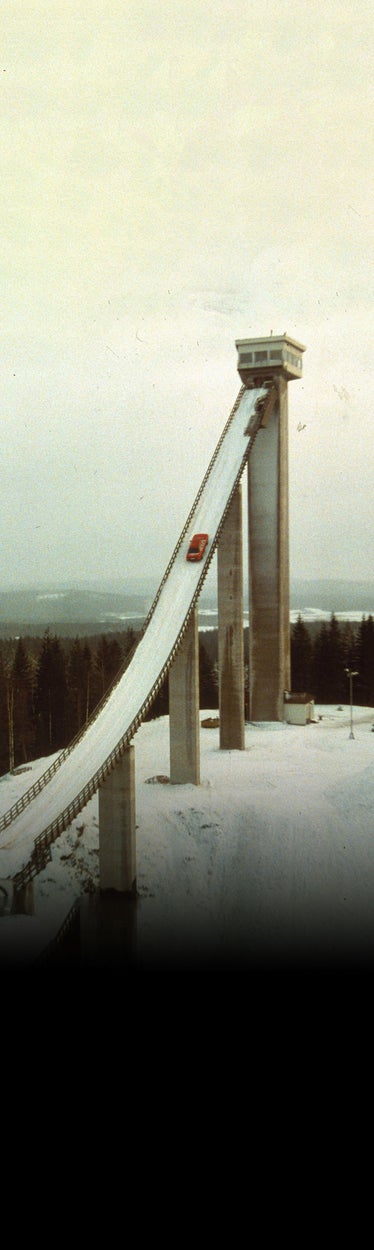 A wide-shot of an early Audi vehicle accelerating up a steep, snowy ramp.