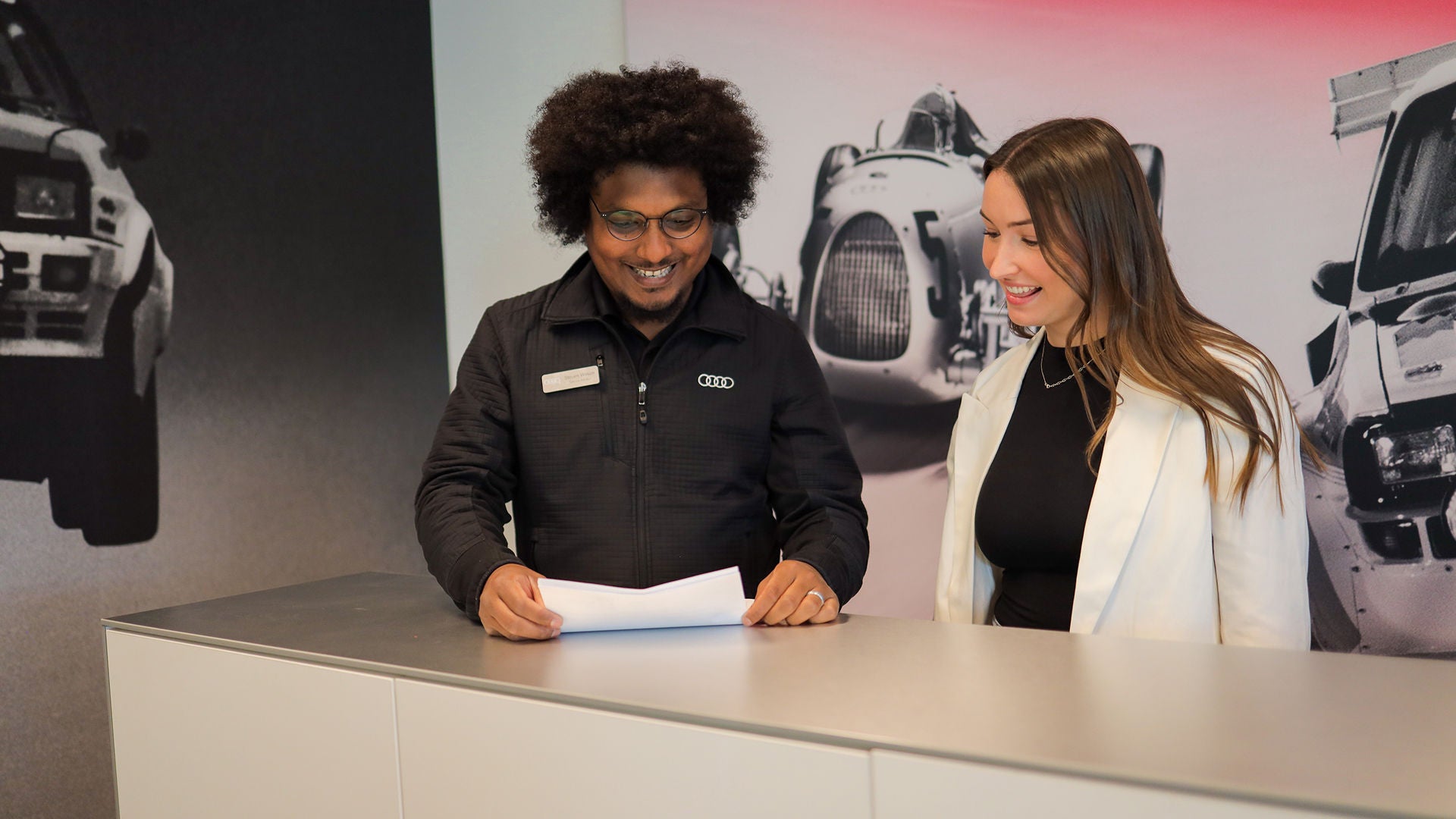Audi sales specialist walking with a customer through the showroom. The sales specialist is handing the customer a key fob for an Audi vehicle. 