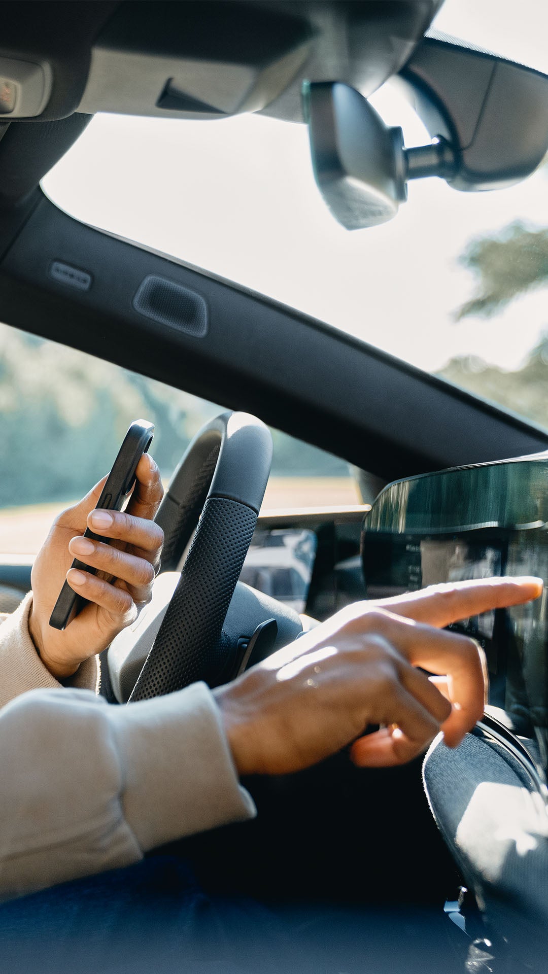 Ein Mann mit beiger Jacke sitzt auf dem Fahrersitz und tippt mit der rechten Hand auf das zentrale digitale Display im Fahrzeug und hält in der linken Hand ein Smartphone.