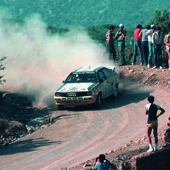 Three-quarter front view of an early Audi racing vehicle accelerating on a dirt road.