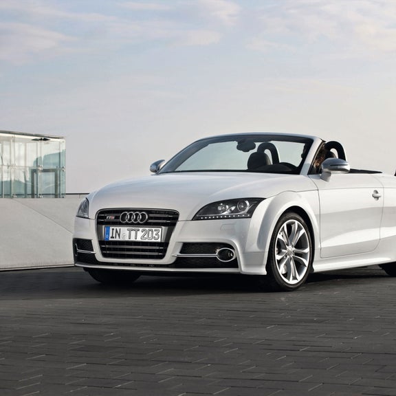 A white Audi TTS Coupe parked on a brick road next to a glass building.