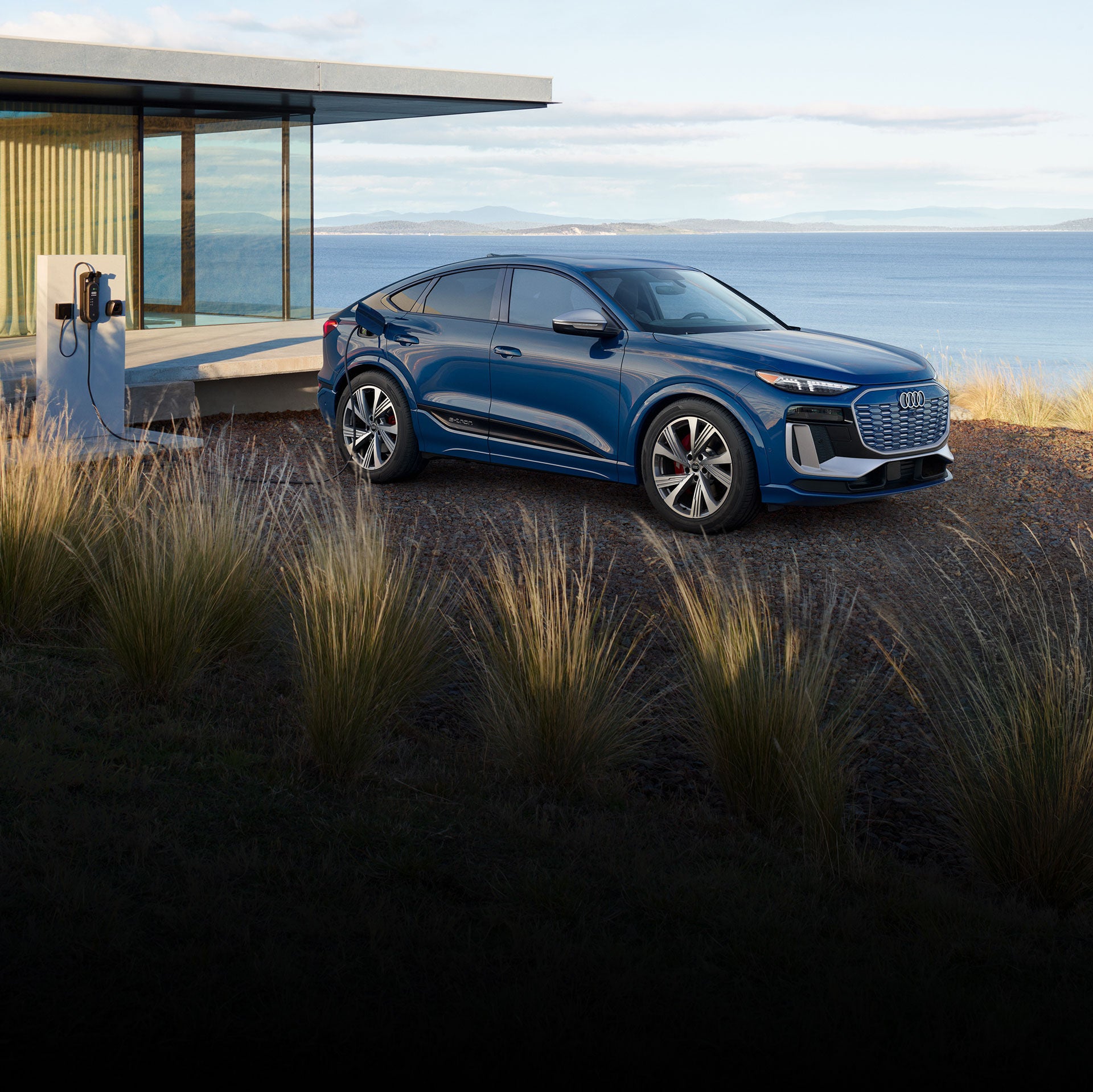 Three-quarter front-view of a grey Audi Q6 Sportback e-tron parked and charging by a waterfront home.