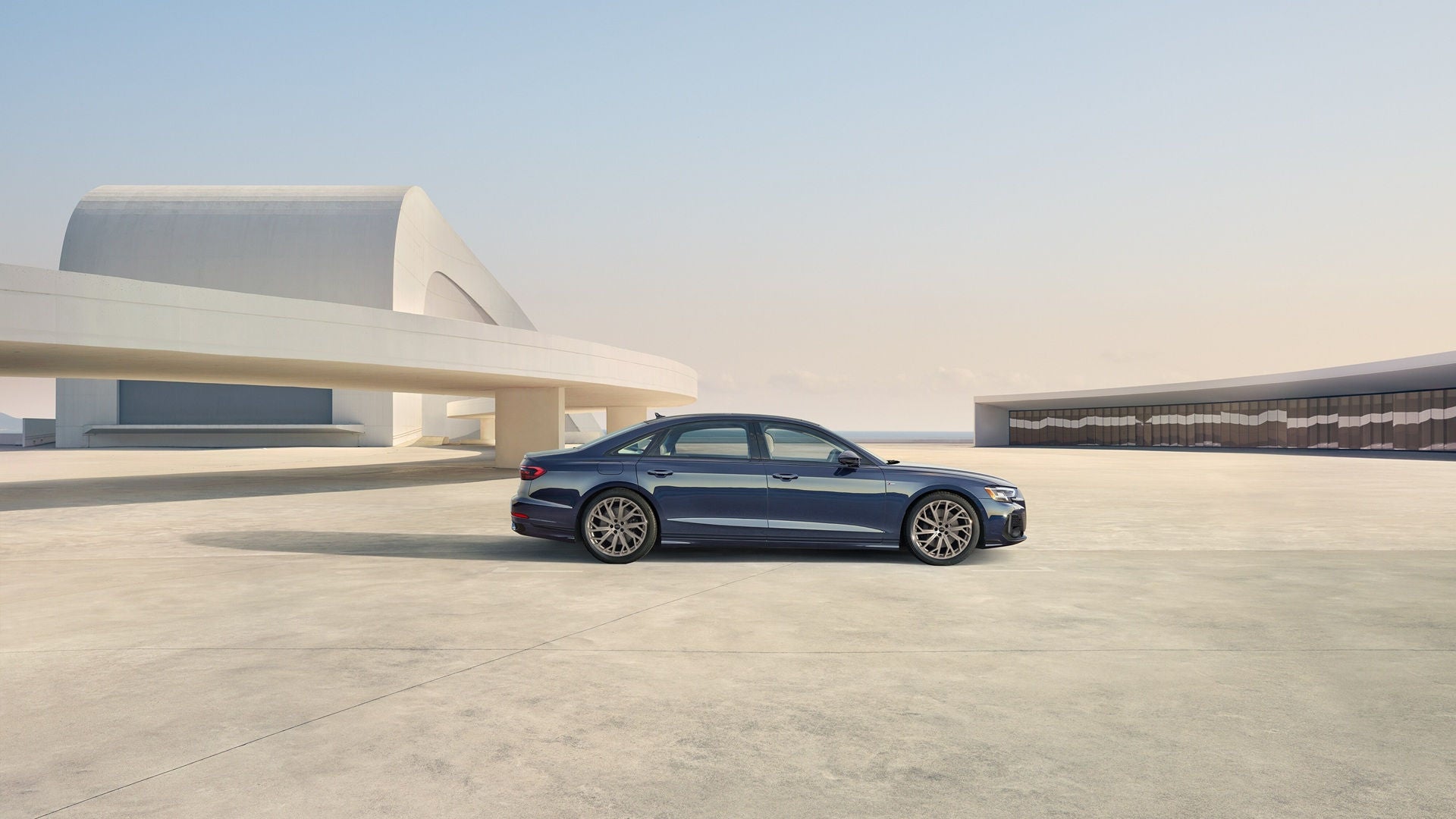 Blue Audi A8 parked on concrete with a modern architect building behind, along with a body of water during the day.  