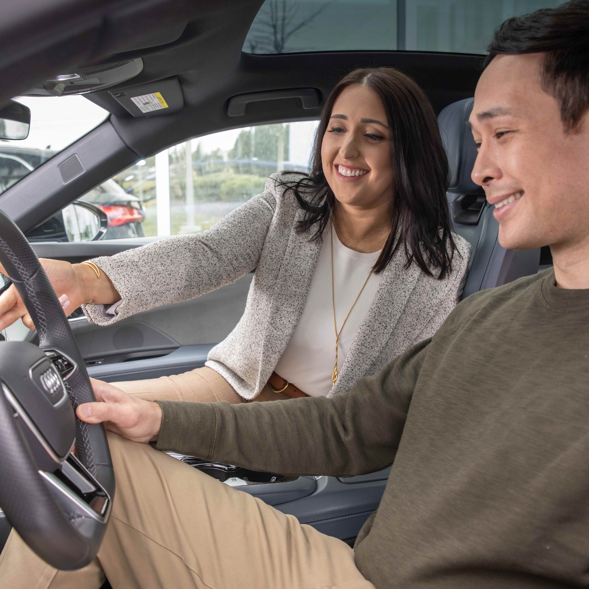 View from the driver's side of a man sitting the driver's seat of an Audi while an Audi Brand Specialist sits in the passenger's seat gesturing towards the steering wheel. Both people are smiling.