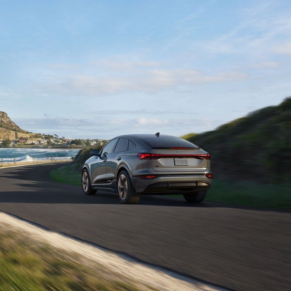 Three-quarter rear view of the Audi Q6 Sportback e-tron accelerating on a seaside road.