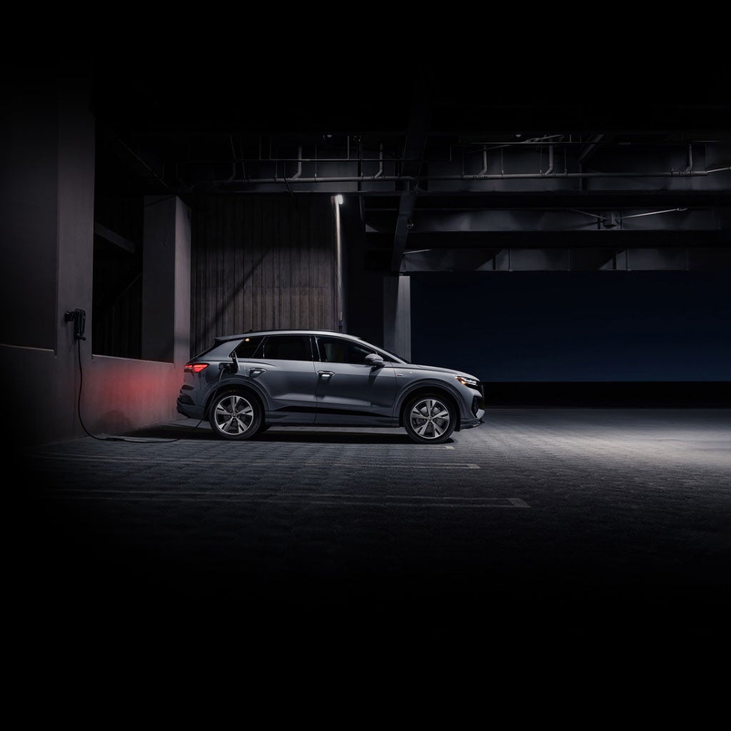 Profil latéral d’un Audi Q4 e-tron argenté en recharge dans un stationnement sombre.